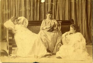 Three college women at the turn of the 20th century, dressed in costumes while one plays mandolin.