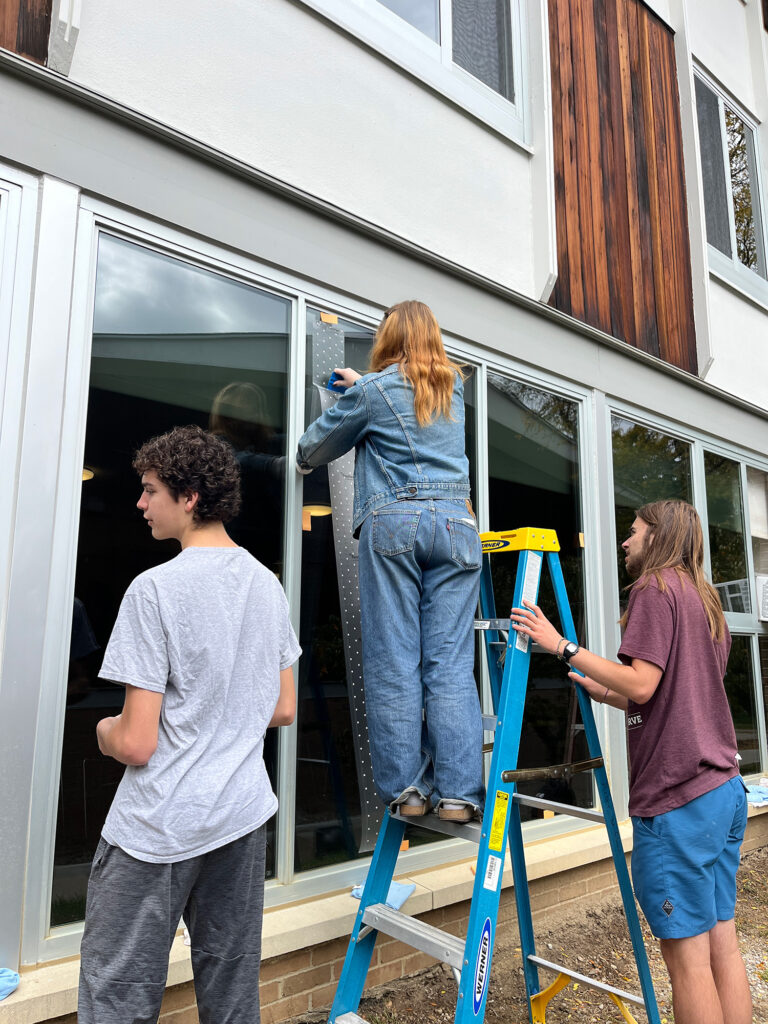 Three people apply patterned film to a window of a building to help deter birds from striking it