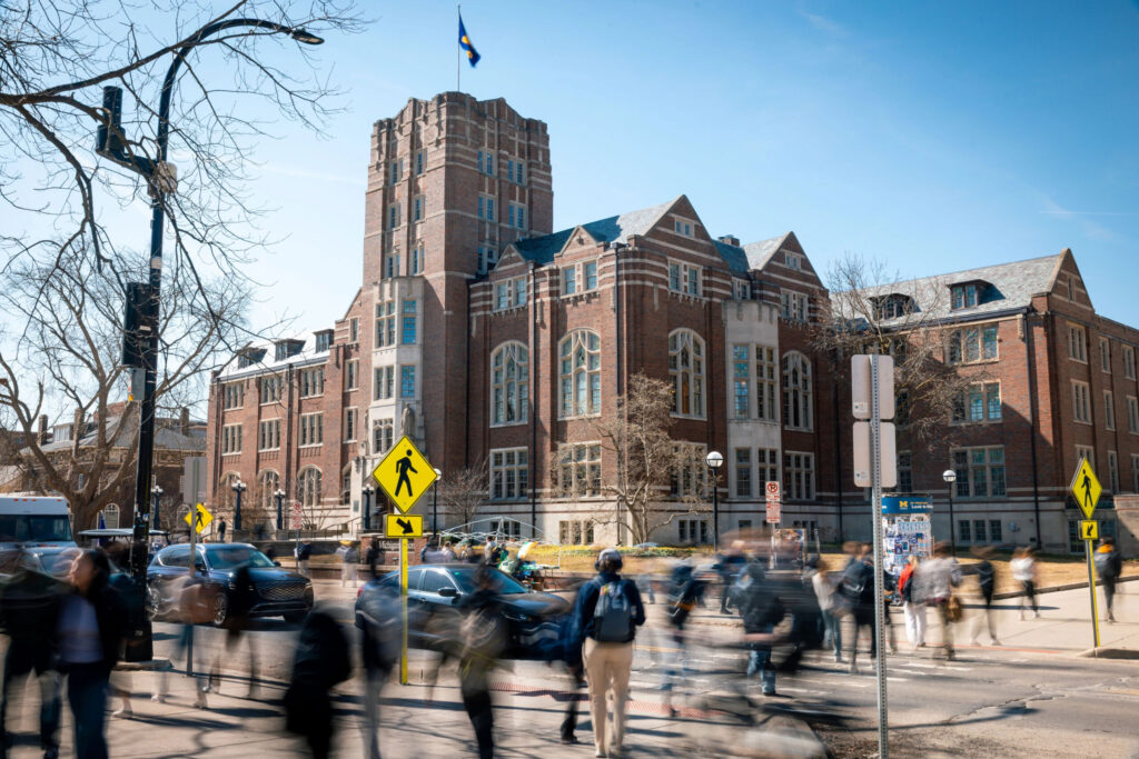 People are blurred walking across the street on a sunny day with a large building in focus in the background