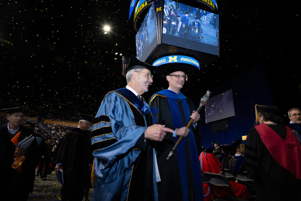 Two men in caps and gowns walk together smiling at a graduation ceremony