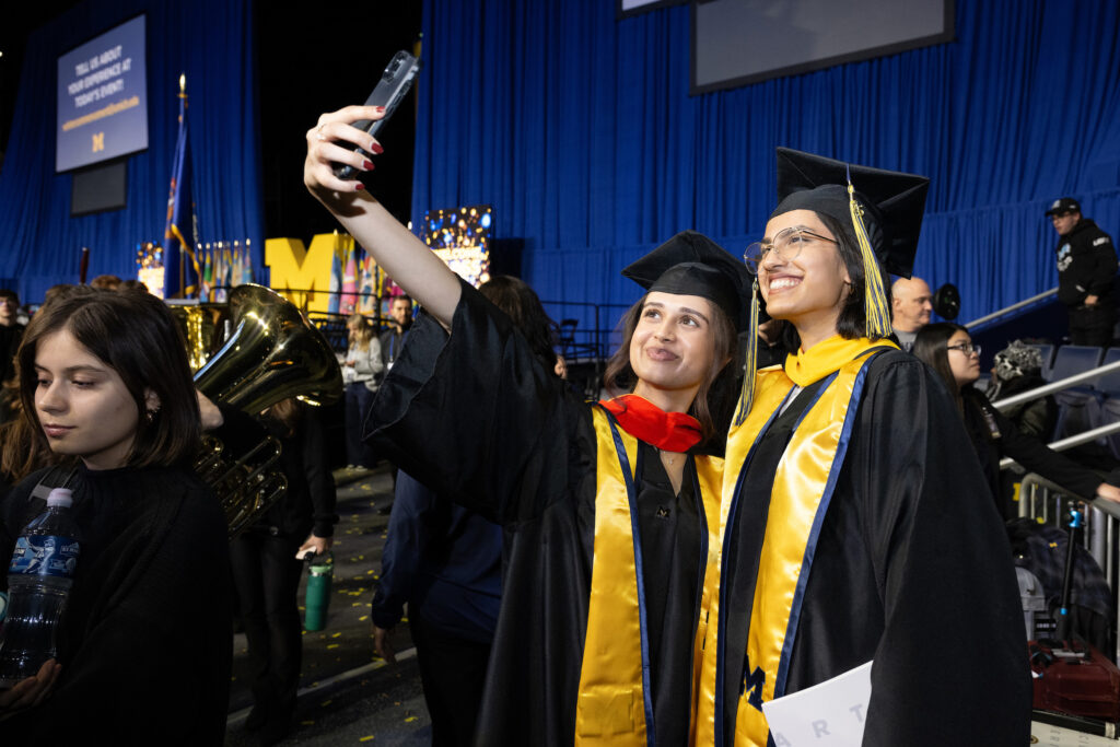 Two women in caps and gowns take a selfie with a cell phone at a graduation ceremony
