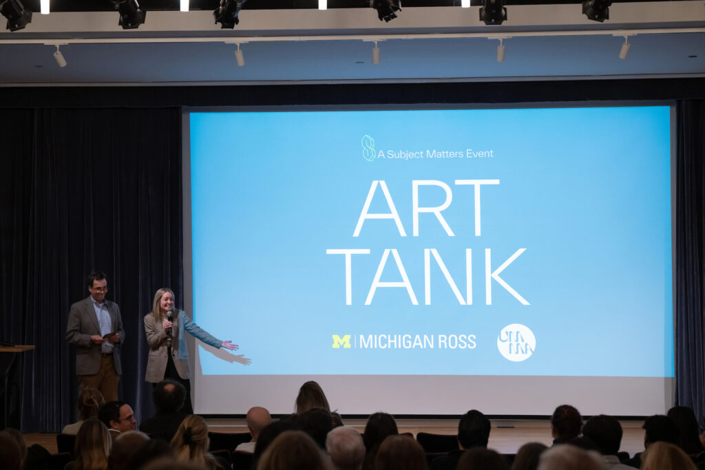 Two people stand on a stage in front of a lrge screen and speak to a crowd