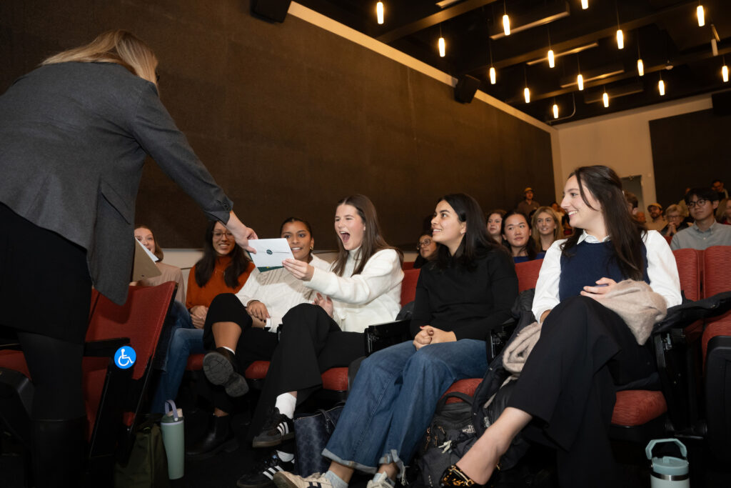 Women sitting in a theater smile as they're presented with an envelope