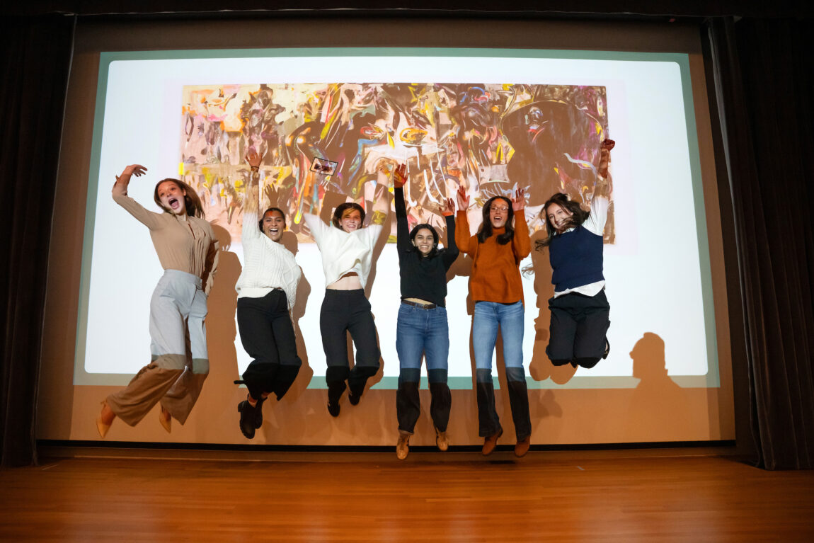 Six people jump in the air in front of a screen displaying art