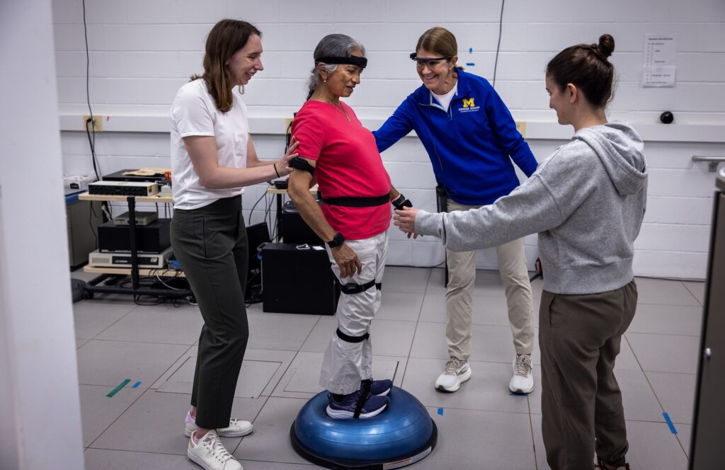 A woman stands on a squishy, plastic dome while wearing black velcro straps around her head, upper arms, wrists, hips, upper thighs, below the knees and feet. Three women surround her with arms extended to catch her if she falls.