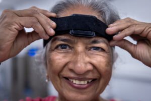 A closeup of a smiling woman’s face. She uses her hands to adjust a black velcro strap positioned around her forehead. The strap has a square, black plastic device in the center.
