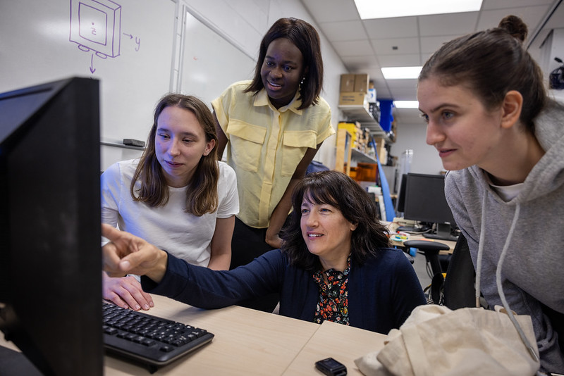 Four women gather together to look at a computer monitor in the foreground. One woman points and speaks while the others look on.