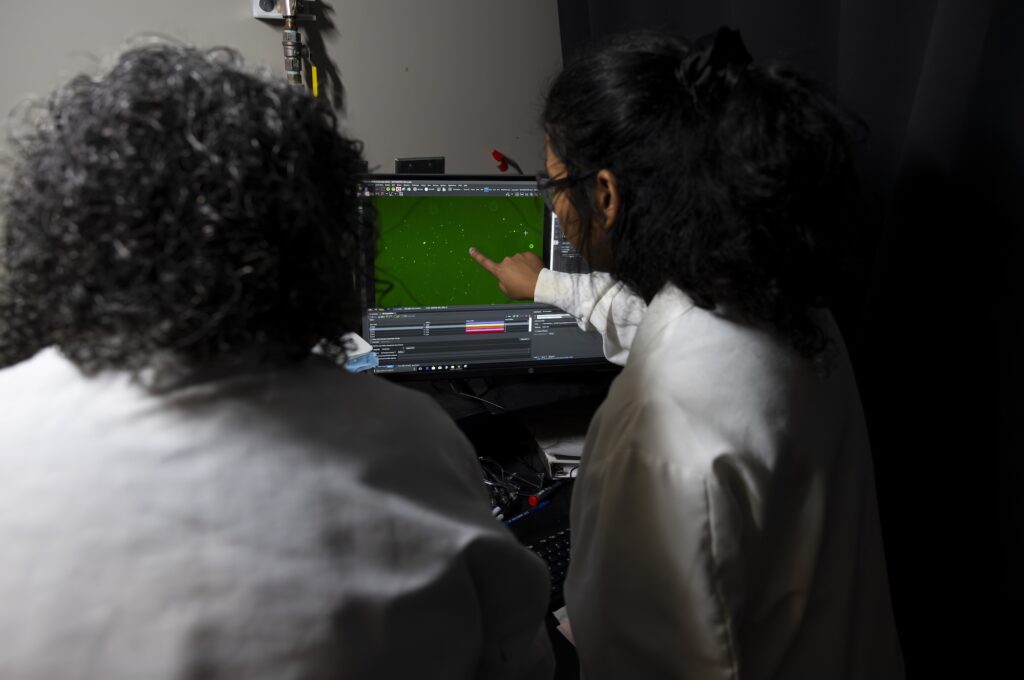 Abha Kumari points at the screen showing the tumor vesicles marked with green fluorescent molecules while Sunitha Nagrath looks on. By counting these vesicles captured from blood samples before and after treatment, the U-M/Northwestern team could predict whether a brain cancer treatment was successful. Image credit: Jeremy Little/Michigan Engineering