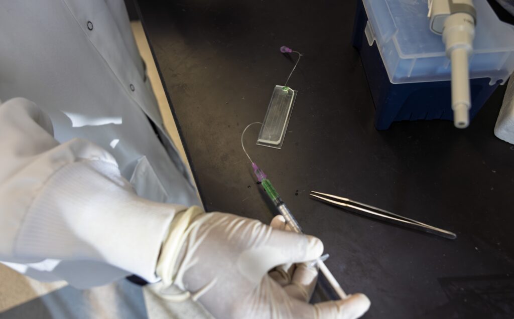 A white gloved hand pushes the plunger on the syringe, which is connected to the chip. The chip is about one inch wide, three inches long, and a quarter of an inch tall.