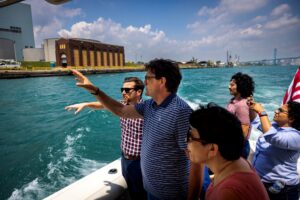 A group of people gesture toward the shoreline from a small motorboat. The city of Detroit is visible on the horizon.
