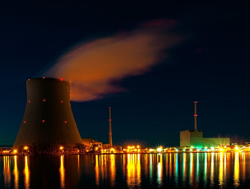 A nighttime view of a power plant with a cooling tower, emitting vapor, reflecting on nearby water.