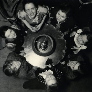 A group of University of Michigan women, circa 1920, are seated around a round table and are photographed from above, looking up at the camera. Black and white.