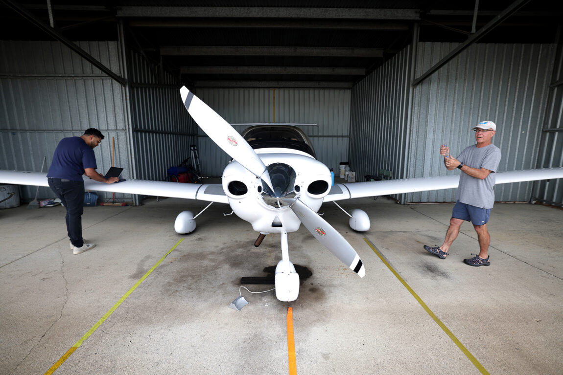 Two men stand near a small, two-person plane inside a hangar. A man on the left of the plane is looking at a laptop on one of the wings.