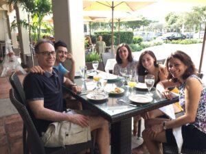 A family of five dines around an outoor table with umbrellas in a seemingly tropical location.