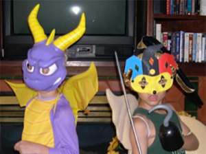 Two kids are dressed up, one likea purple dragon type creature and another like a colorful jester.