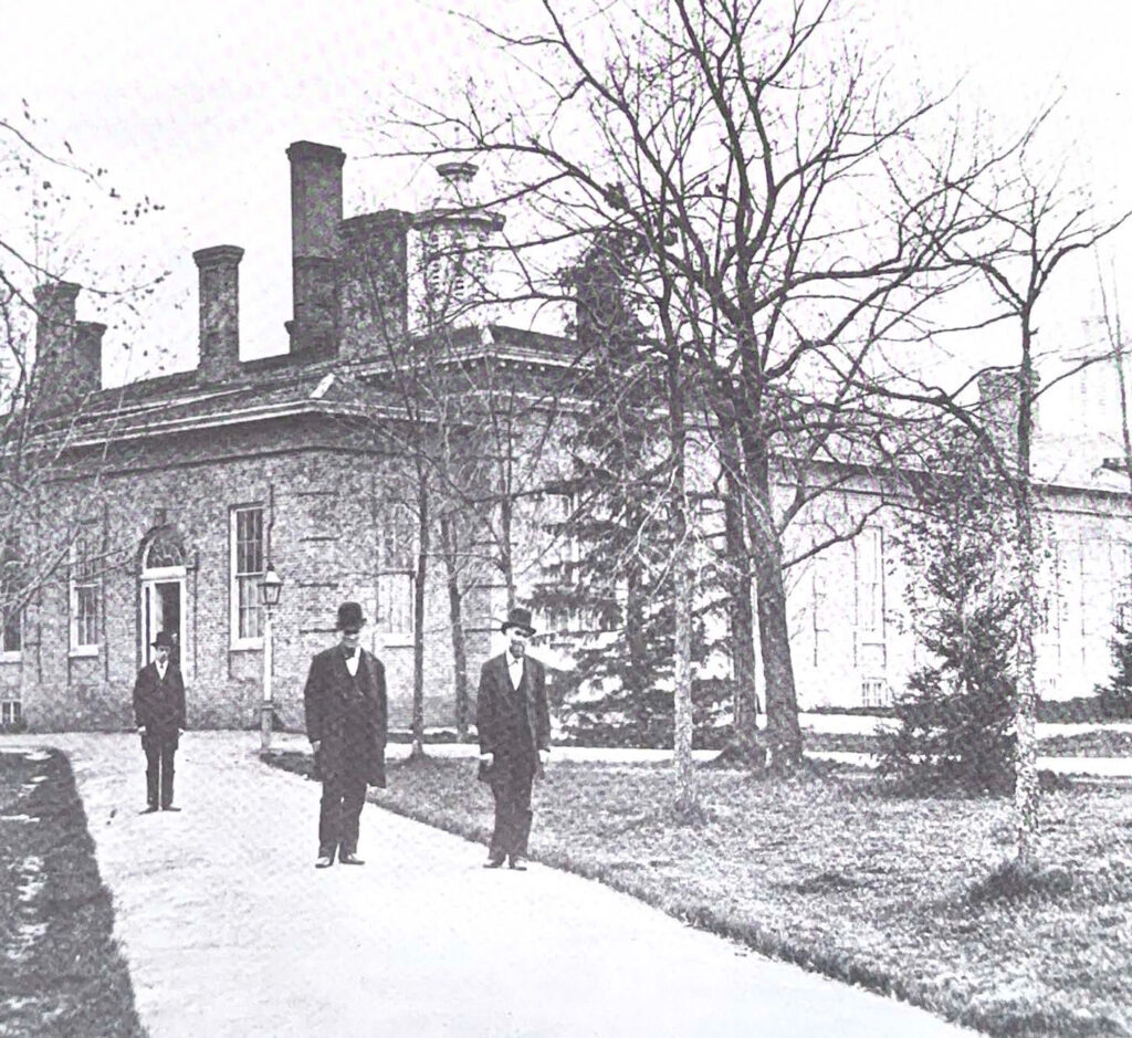Three men in suits and top hats walk on a sidewalk in front of an old building