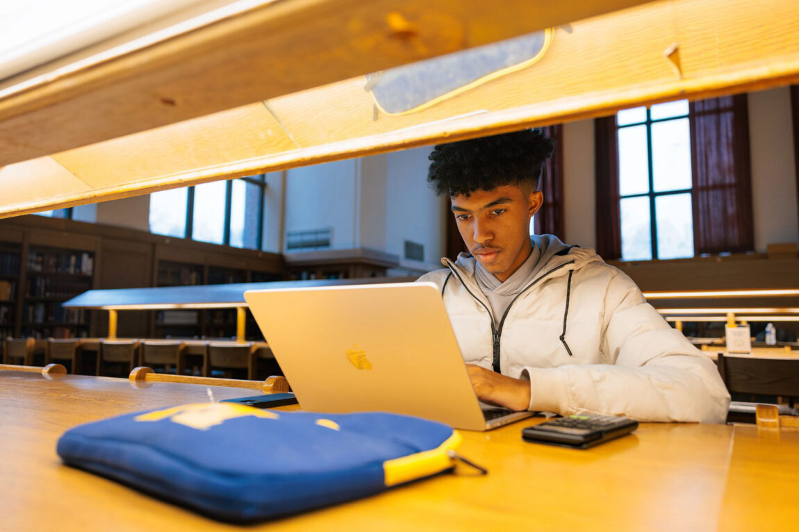 A student in a gray sweatshirt studies on their laptop at one of the long tables in the Hatcher Graduate Library reference room.
