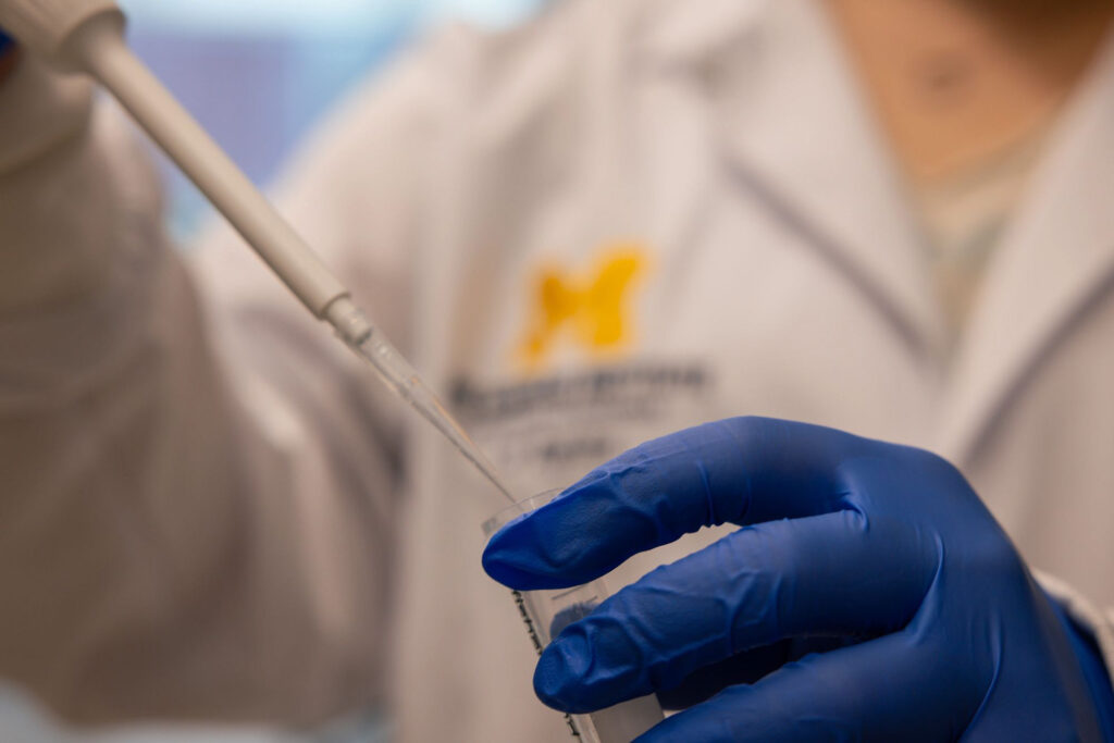 A student in a lab coat with the Block M embroidered on the chest pipes liquid into a beaker.
