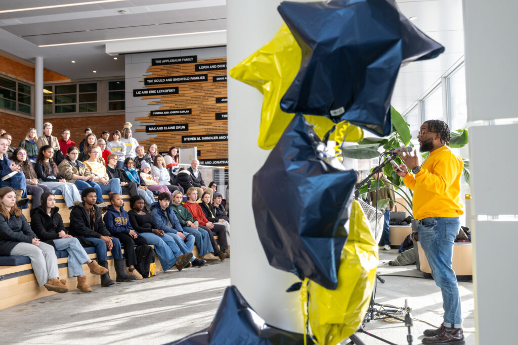 A man standing at a microphone speaks to a crowd of people with blue and yellow star-shaped balloons in the foreground