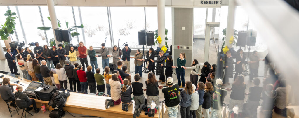An overhead view of dozens of people in a room forming a circle with their arms crossed in front of them, joining hands with the people next to them