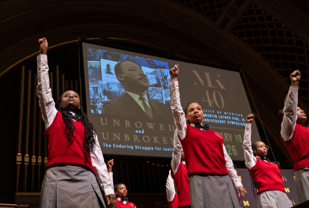 Several young people dressed in red vests and gray skirts or slacks raise one arm with fist clenched on a stage