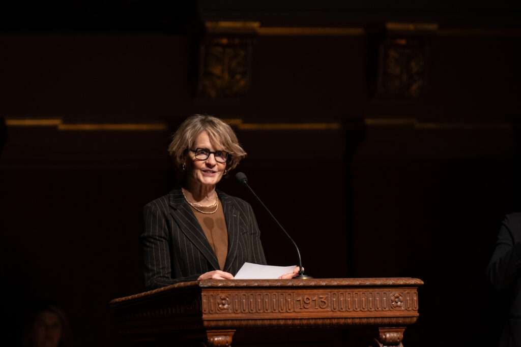 A woman wearing glasses holds a piece of paper and speaks at a podium