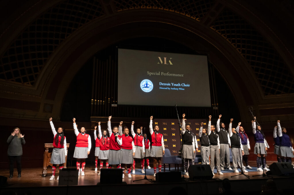 Several young people dressed in red or black vests with gray skirts or slacks stand on a stage with one arm raised and fist clenched
