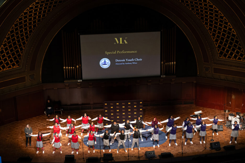 Several young people wearing red, black or blue vests and gray skirts or slacks stand on a stage with their arms outstretched