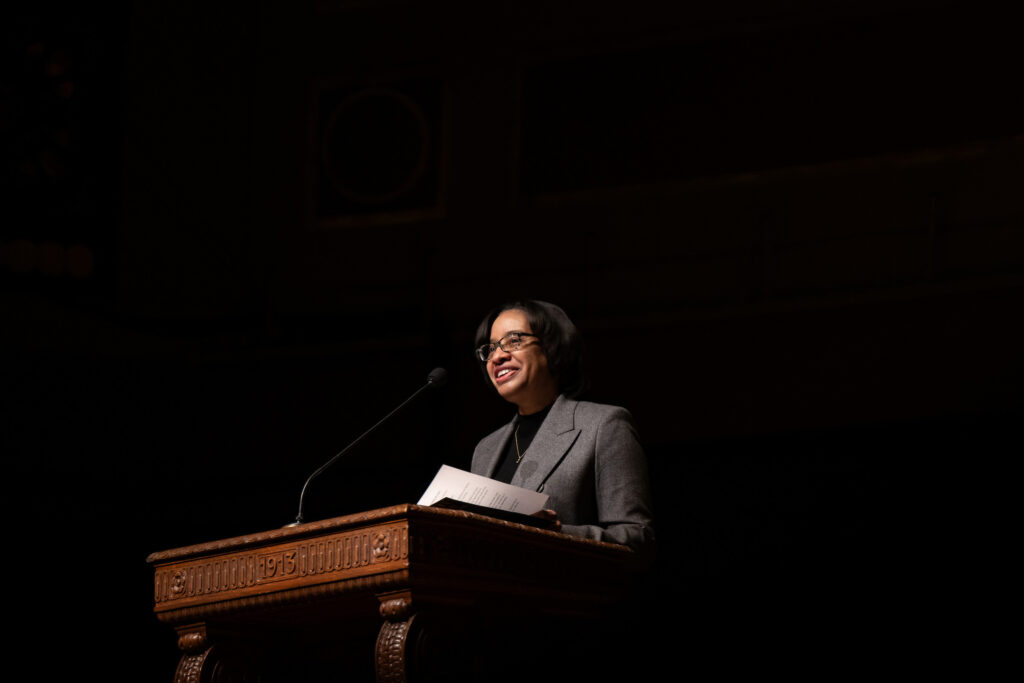 A woman stands at a lectern and smiles as she speaks to a crowd
