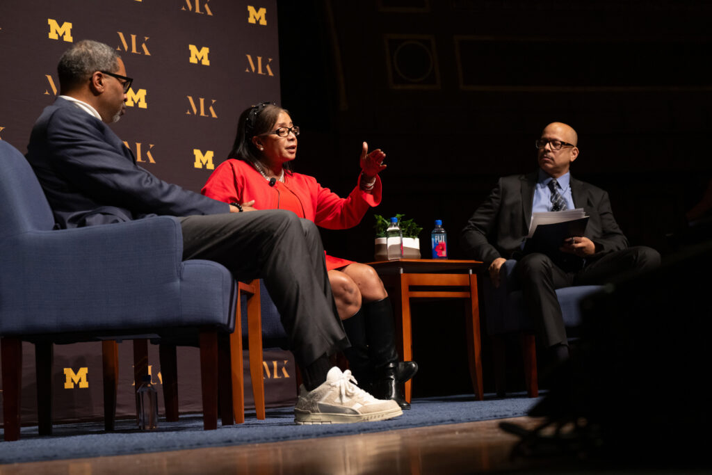 Two men and a woman sit in chairs on a stage and speak to a crowd