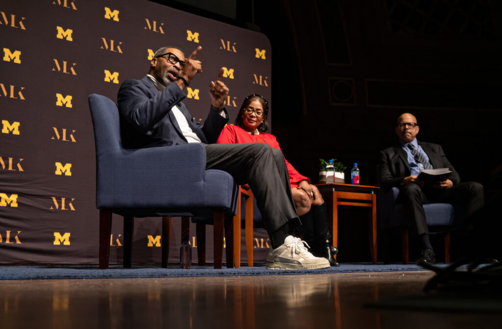 Two men and a woman sit in chairs on a stage and speak to a crowd