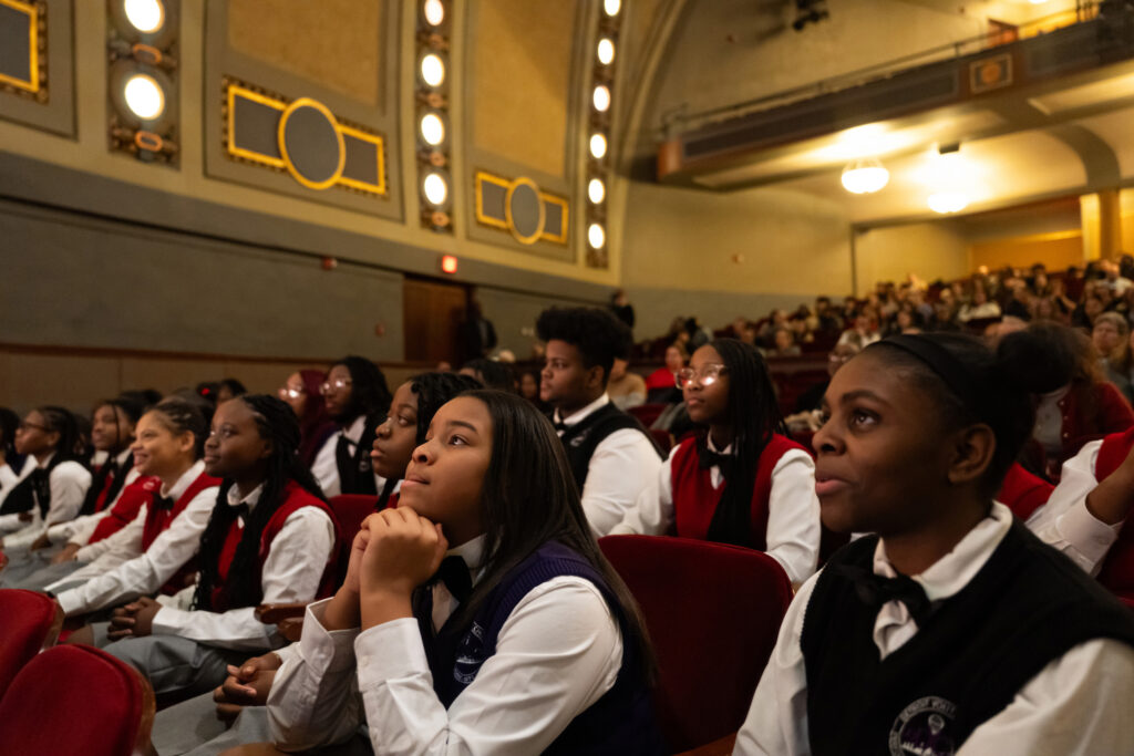 Young people seated in chairs in an auditorium watch a performer on stage