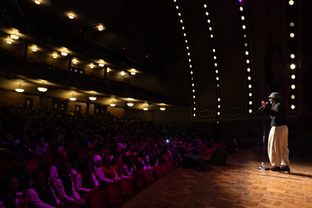 A person performs on a stage as people seated in an auditorium listen