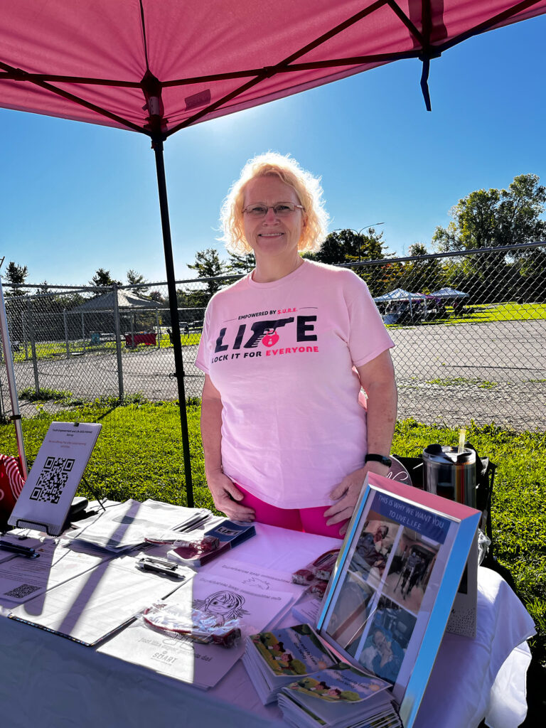 A woman wearing a pink shirt stands underneath a canopy at a table on a sunny day